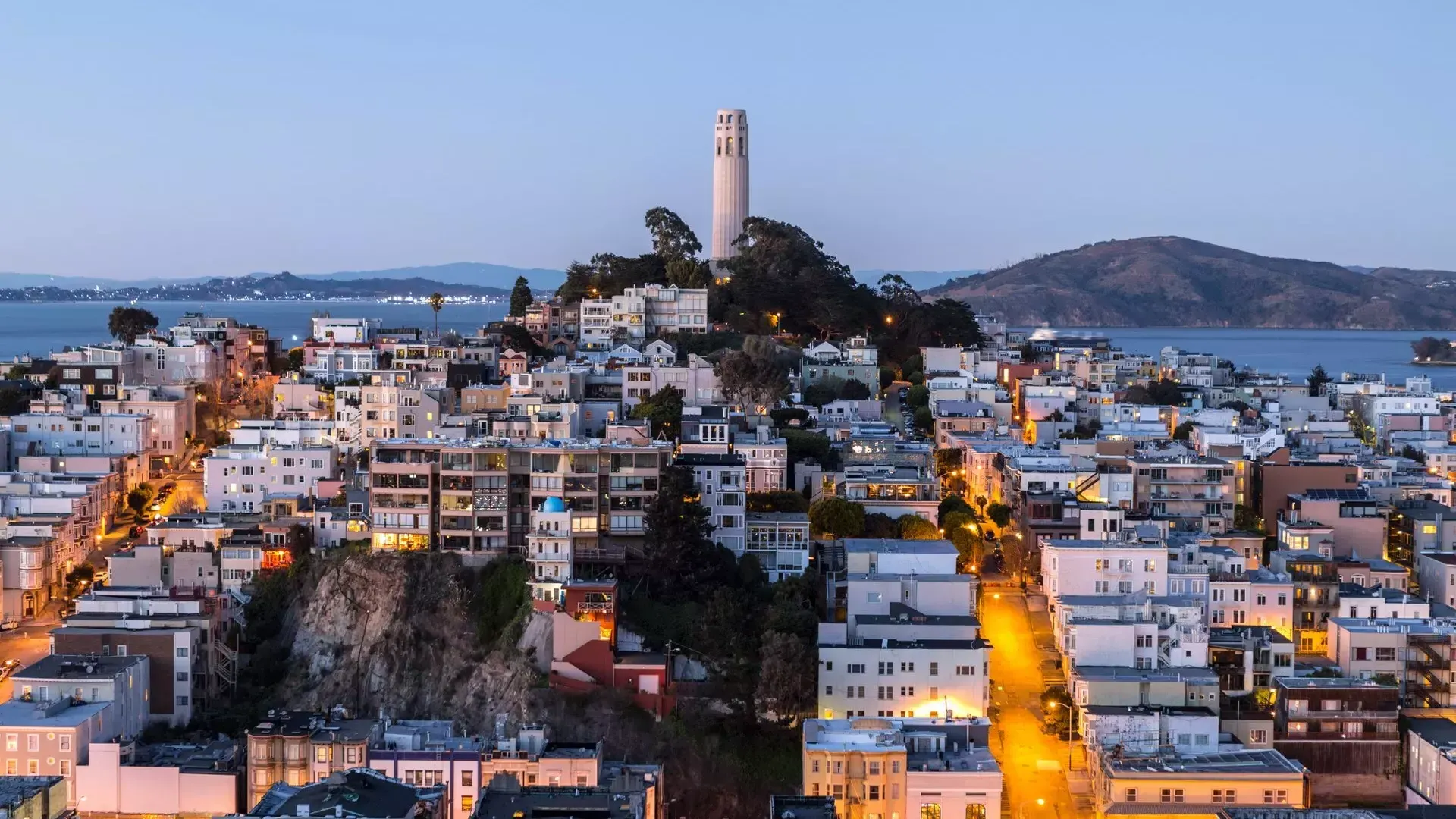 Coit Tower de San Francisco au crépuscule, avec les rues éclairées devant elle et la baie de San Francisco derrière elle.