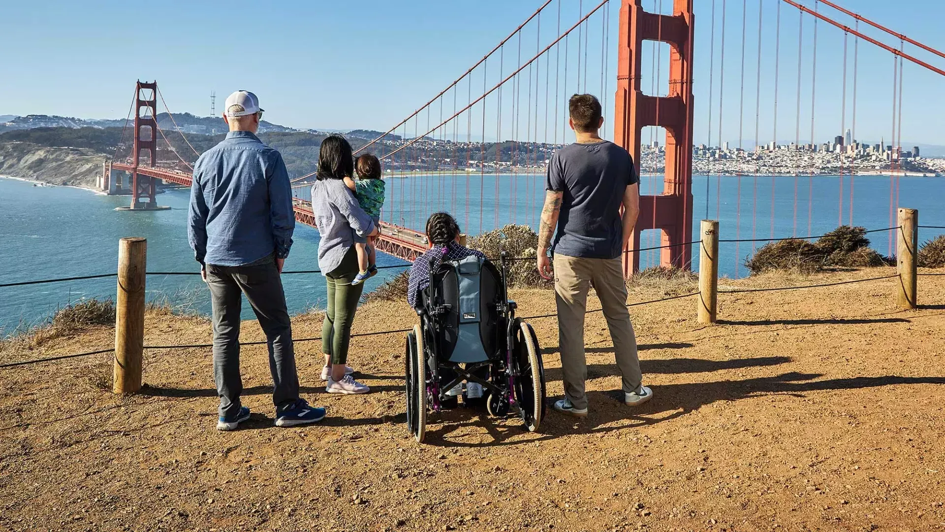 Um grupo de pessoas, incluindo uma pessoa em uma cadeira de rodas, é visto de trás enquanto olham para a Golden Gate Bridge de Marin Headlands.