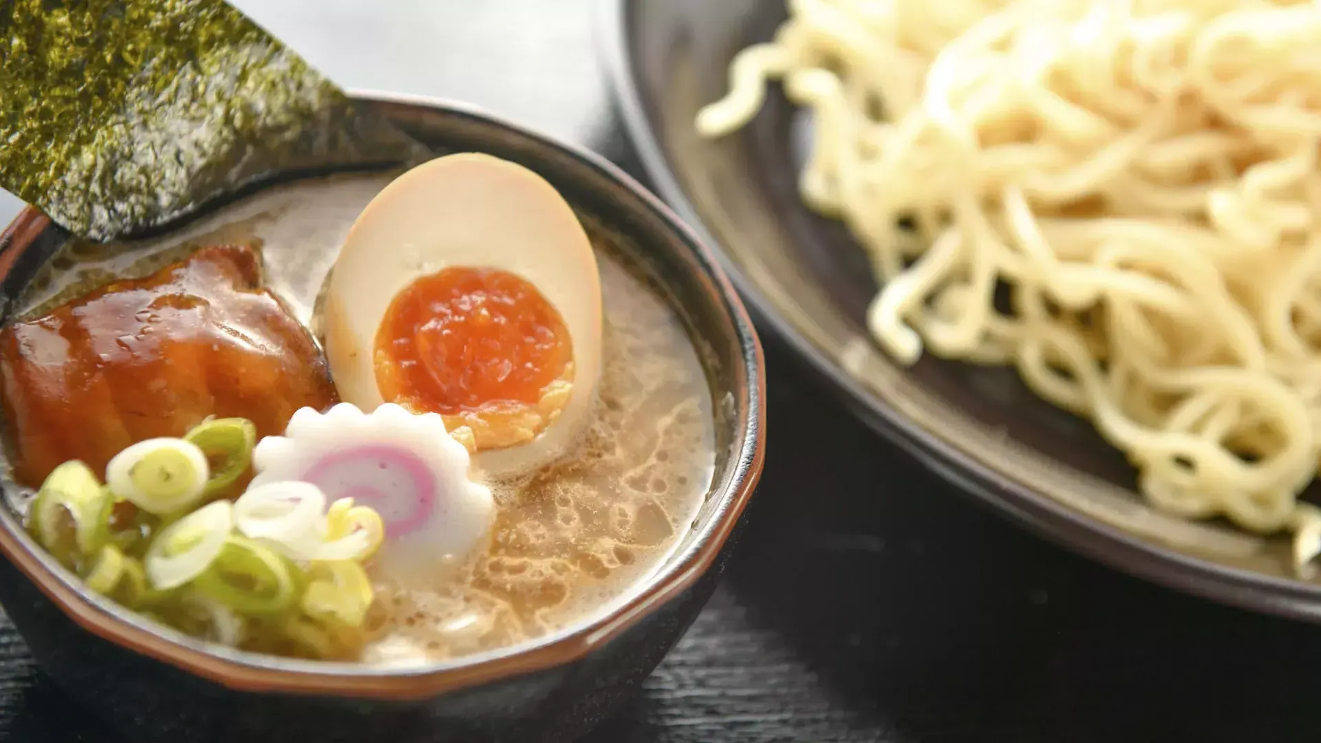 Close-up de uma tigela de macarrão e uma tigela de sopa de ramen com um ovo escalfado, cortado ao meio.